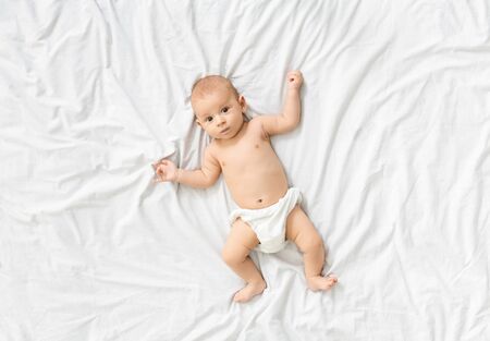 Calm curious infant in diaper lying on bed at home, looking at camera, top view, copy spaceの写真素材