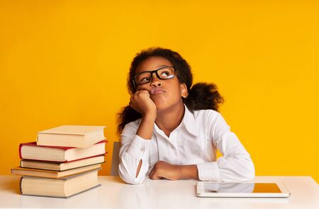 Bored Black School Girl Sitting Between Tablet Computer And Book Stack On Yellow Background In Studio. Boring Homework Conceptの写真素材