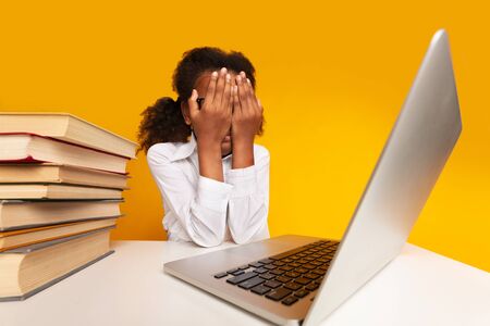 Overworking Black Schoolgirl Covering Face With Hands Sitting At Laptop On Yellow Background In Studio. School Overload Conceptの写真素材