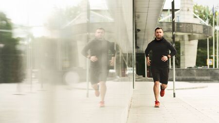 Handsome male athlete jogging near urban building with mirror windows, panorama with copy space