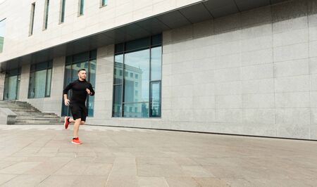 Serious millennial man jogging outdoors against urban building, training endurance. Panorama with...