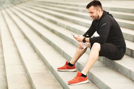 Man texting on his mobile phone during workout in city, having rest, sitting on steps, side view...