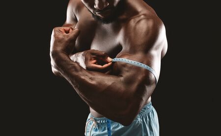 Muscular african american bodybuilder measuring himself biceps with tape measure, cropped, black studio backgroundの写真素材