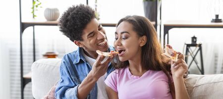 Teenage couple having fun at home, black guy feeding his girlfriend with pizza. Panoramaの写真素材