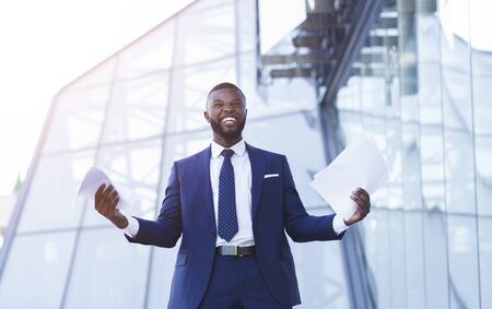 Business Success. Excited Black Businessman Gesturing Yes Holding Papers Outdoor Against Modern...