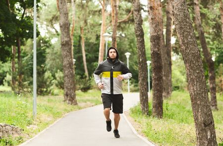Young sporty guy in windbreaker jogging on valley in autumn park, training endurance for marathon