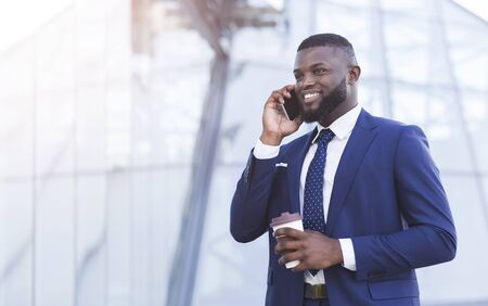 Smiling Afro Business Guy Talking On Mobile Phone Holding Coffee Cup Walking Outdoor Next To Modern...