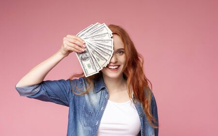 Cashback. Happy redhead girl covering her face with a fan of dollars, showing her profitの写真素材