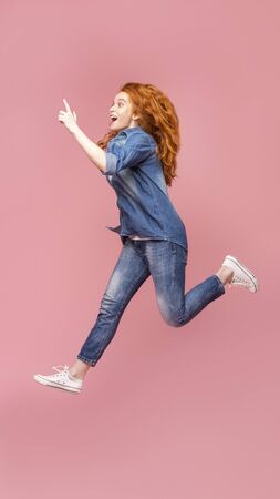 Look there. Portrait of beautiful redhead girl jumping and pointing finger aside on pink studio background, full-length shot with free spaceの写真素材