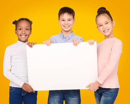 Happy diverse kids showing blank placard board for text over yellow backgroundの写真素材