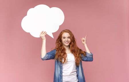Inspired ginger girl holding blank speech bubble, having idea and pointing finger up over pink...