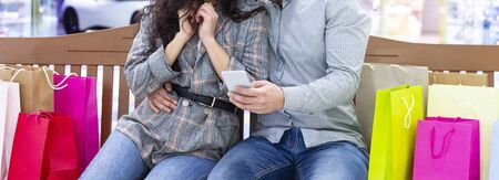 Online purchases. Couple resting in shopping mall on bench, using phone, cropの写真素材