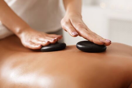 Masseur doing back hot stone massage to woman in spa salon, closeup