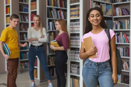Cheerful black girl posing in front of friends, staying next to bookshelves in campus libraryの写真素材