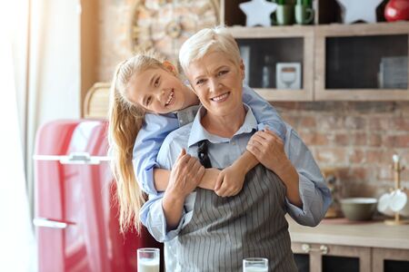 Cheerful little granddaughter hugging her happy grandmother at kitchen at home, copy spaceの写真素材