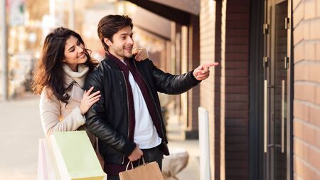Happy young couple shopping together, man pointing at shop window, showing clothes to wifeの写真素材