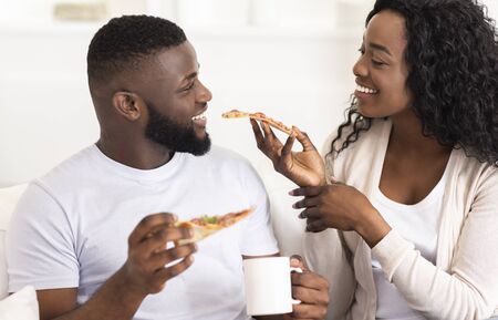 Lovely black millennial couple sharing pizza and drinking coffee on date at homeの写真素材