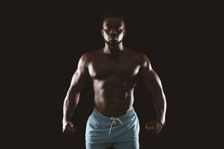 Handsome african bodybuilder showing muscular body, stayng in shadow, black backgroundの写真素材