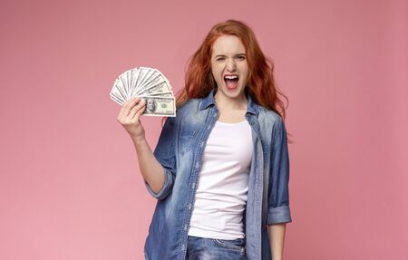 Yes, I did it. Happy girl with red hair holding bunch of money banknotes and celebrating profit,...