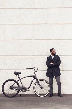 Young african man with bicycle standing over white wall outdoors looking aside, free space