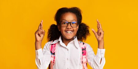 Cheerful Black Schoolgirl Keeping Fingers Crossed For Luck In First School Day. Panorama, Yellow Background, Studio Shotの写真素材