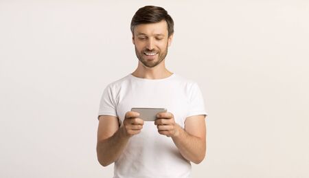 Cheerful Man Using Mobile Phone Playing Games Or Texting Over White Studio Background. Copy Space