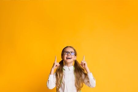 Look Up. Excited First-Grade School Girl Pointing Fingers Upward At Free Space For Text Over Yellow...