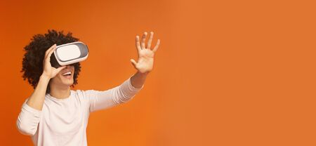 Emotional millennial guy enjoying virtual reality glasses, pushing invisible button, orange panorama...