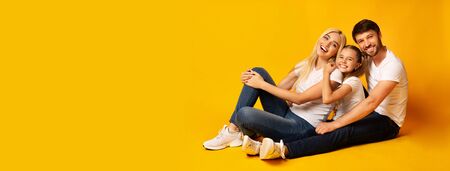 Positive Family Of Three Hugging Relaxing Together Sitting On Floor Over Yellow Studio Background. Copy Space, Panoramaの写真素材