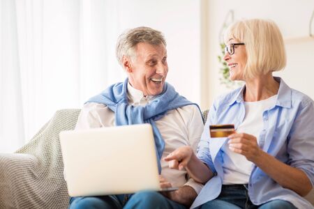 Happy mature couple making online purchases on laptop, woman holding credit card, resting at home