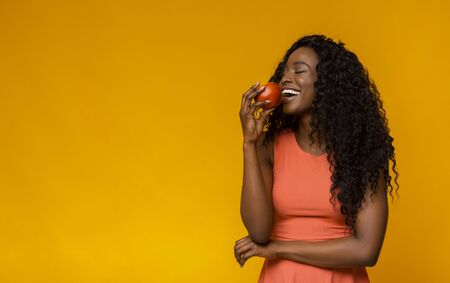 Happy African American Lady Enjoying Red Apple, yellow studio background, copy spaceの写真素材