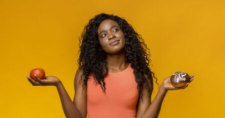 Healthy lifestyle or nutrition concept with young african woman holding in hands apple and donut,...