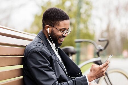 Happy afro man sitting in park and listening music with earphones, having break outdoors