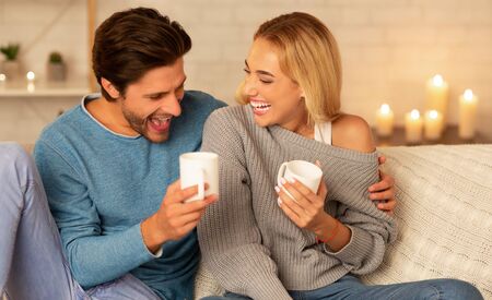 Stay-At-Home Date. Cheerful Couple Laughing Sitting On Couch Drinking Coffee Indoor At Night....