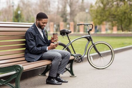 Coffee break. Happy businessman using phone, resting next to bike in parkの写真素材