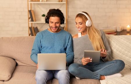 Modern Lifestyle. Smiling Couple Using Laptop And Tablet Computers Wearing Headphones Sitting On...