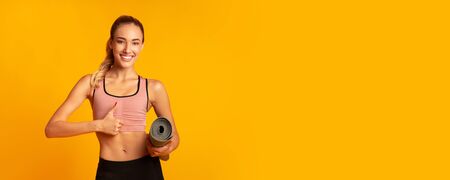 Cool Fitness Workout. Positive Girl Holding Rolling Mat Gesturing Thumbs-Up On Yellow Studio Background. Panorama, Empty Spaceの写真素材
