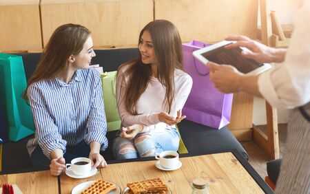 Two attractive girls discussing what to order in cafe after shopping day, waiter with tablet, blank...