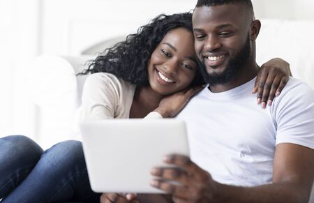 Happy black couple browsing social media on digital tablet sitting together on couch at home, copy spaceの写真素材