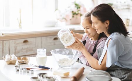 Mother and daughter preparing dough for bake cookies, adding flour to bowl, having fun in kitchen...
