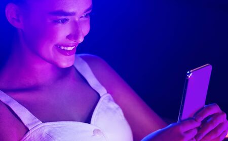 Young woman using smartphone in room with blue and pink neon lightsの写真素材