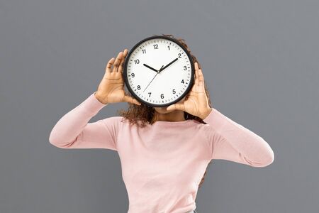 Time management concept. Young mixed race woman covering her face with big watch on grey background