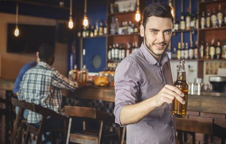 Handsome young guy raising beer bottle and smiling, spending time with friends at pub, panorama with copy spaceの写真素材