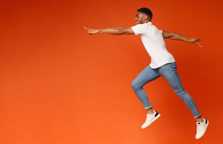 On way to aim. Happy african guy jumping in air, reaching hand aside to free space, orange backgroundの写真素材