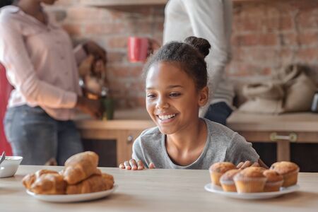 Charming little african girl looking with joy at sweets while parents busy, kitchen interior, copy spaceの写真素材
