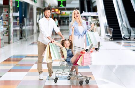 Happy Parents Riding Little Daughter In Shopping Cart Carrying Colorful Bags Smiling At Camera In Mall.の写真素材