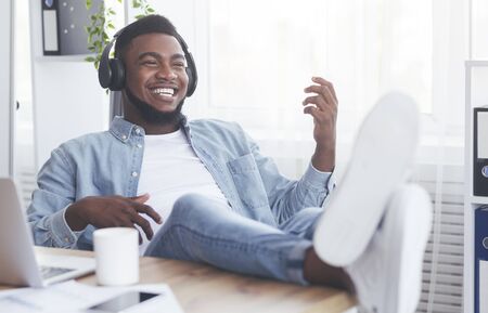 Break at work. Cheerful african american employee listening music in headphones and playing virtual guitar, relaxing at workplaceの写真素材