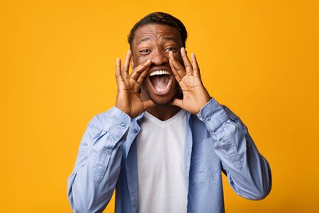 Hey. Emotional Black Guy Screaming Holding Hands Near Mouth Looking At Camera Standing Over Yellow Background. Studio Shotの写真素材