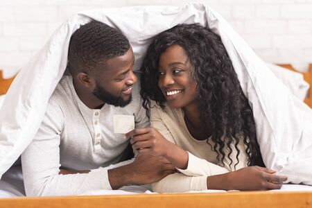 Safe sex. Young african american couple holding condom, lying on bed under white blanket, looking at each other with love and careの写真素材