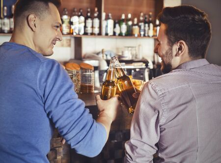 Two cheerful guys having fun together in pub, clinking beer bottles, empty space, back viewの写真素材